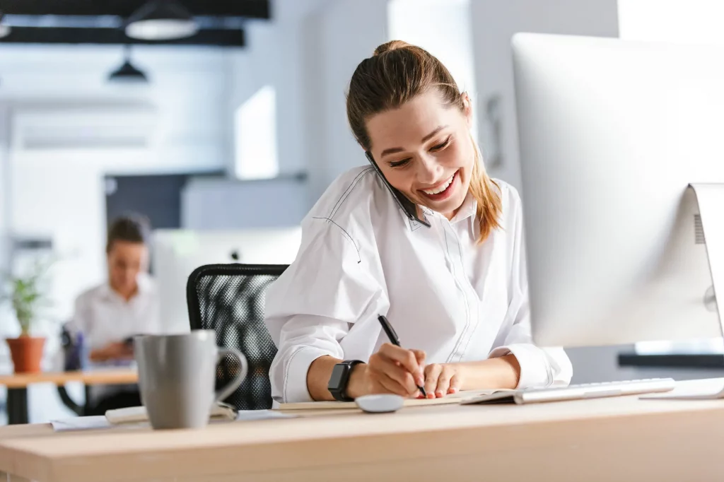 Eine Frau telefoniert lächelnd im Büro und schreibt währenddessen Notizen.