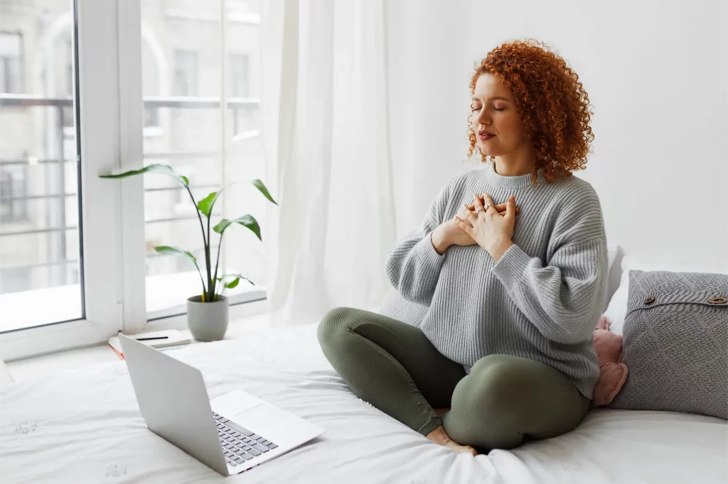 Eine Frau nimmt online mit dem Laptop an einem Webinar oder Workshop teil und macht eine Atem-/Entspannungsübung.