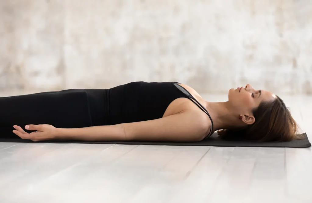 Eine Frau von der Seite in der Yoga-Entspannungsposition auf einer Matte in einem hellen Raum.