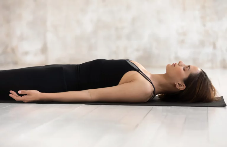 Eine Frau von der Seite in der Yoga-Entspannungsposition auf einer Matte in einem hellen Raum.