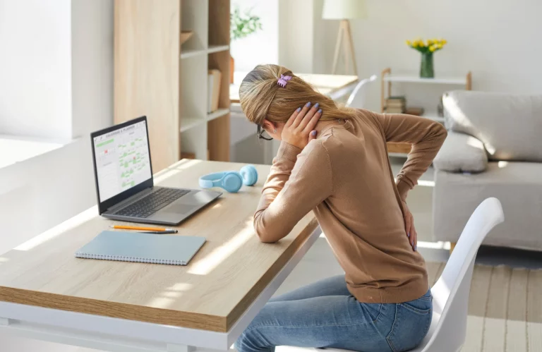 Frau am Schreibtisch mit Rücken- und Nackenschmerzen durch langes Sitzen am Computer.