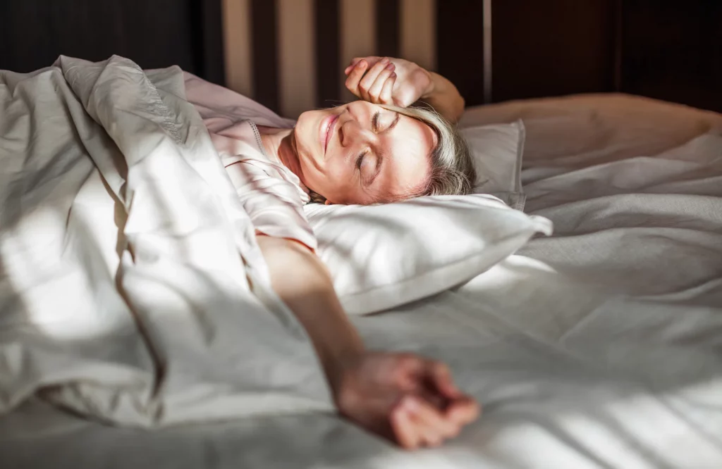 Eine lächelnde Frau mittleren Alters liegt im Bett und räkelt sich entspannt.