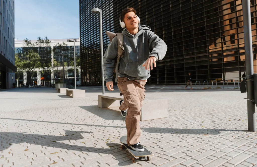 Ein junger Mann fährt lächelnd Skateboard und hört dabei Musik über Kopfhörer.