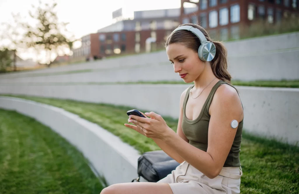 Eine Frau mit Kopfhörern sitzt im Freien und schaut sich Inhalte auf ihrem Smartphone an. Sie trägt am Arm ihr Blutzuckermessgerät.
