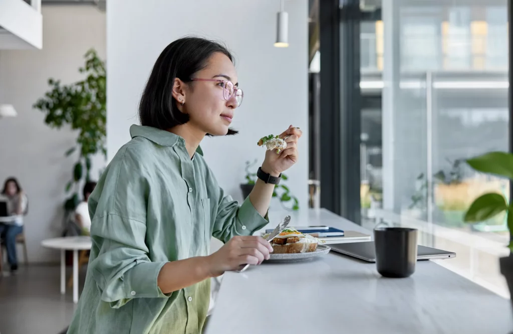 Eine Frau sitzt mit Brille am Fenster einer gemütlichen Cafeteria und isst zu Mittag.