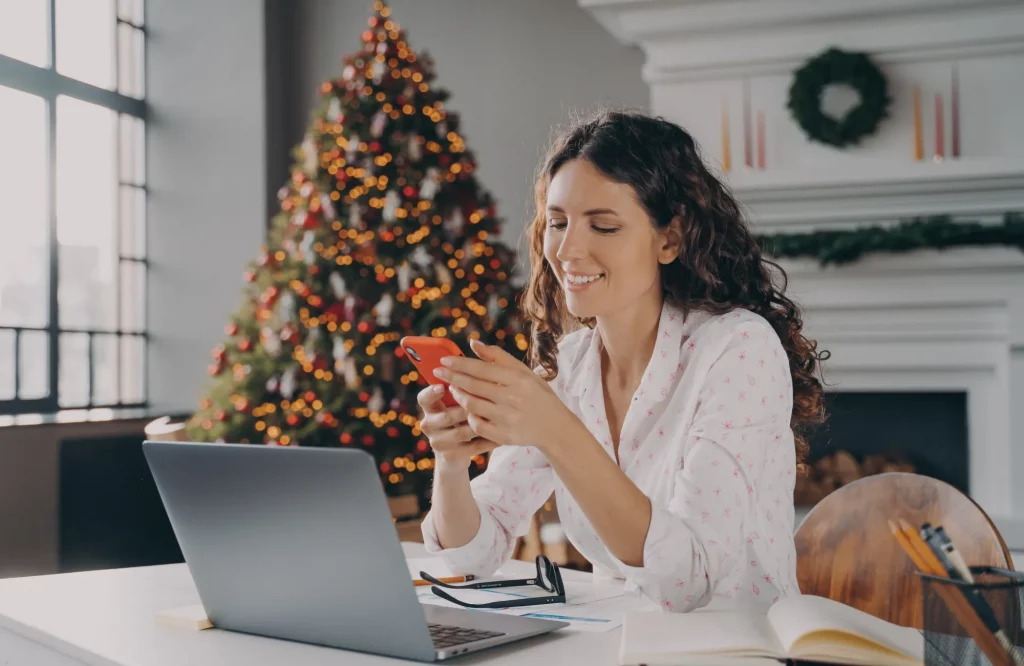 Eine Frau sitzt im weihnachtlichen Büro und schaut lächelnd auf ihr Smartphone.