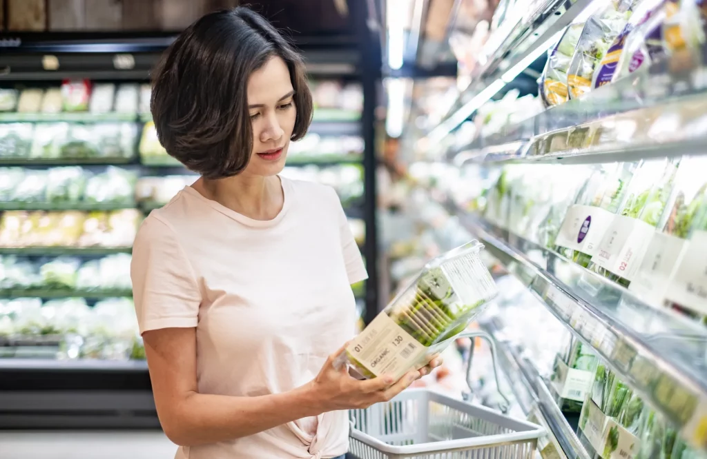 Eine Frau geht mit einem Einkaufskorb durch den Supermarkt und wählt grünen Salat aus dem Regal aus.