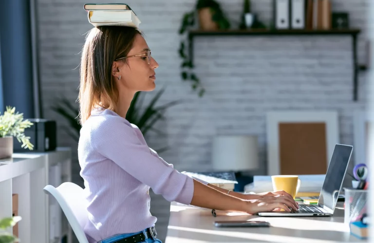Frau sitzt am Schreibtisch und balanciert Bücher auf ihrem Kopf, während sie am Computer arbeitet.