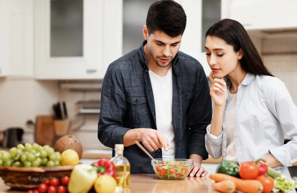 Ein junges Paar bereitet gemeinsam einen Gemüsesalat zu; ein Mann zeigt auf eine Schüssel und unterhält sich über die Zutaten.