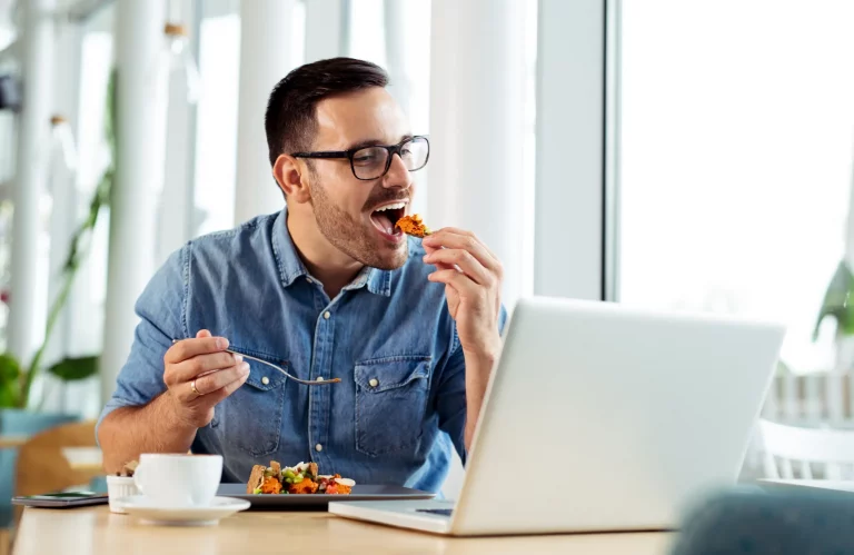 Lächelnder Geschäftsmann genießt ein Mittagessen während der Arbeit vor einem Laptop.