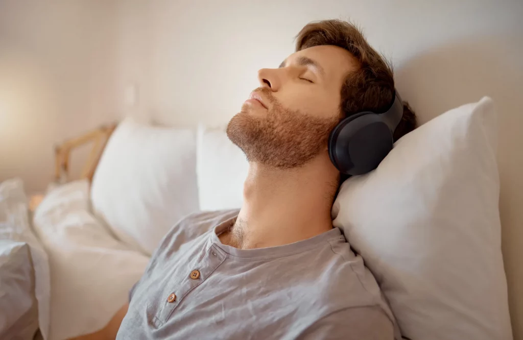 Ein entspannter Mann liegt mit Kopfhörern auf seinem Bett und hört Musik oder einen Podcast.