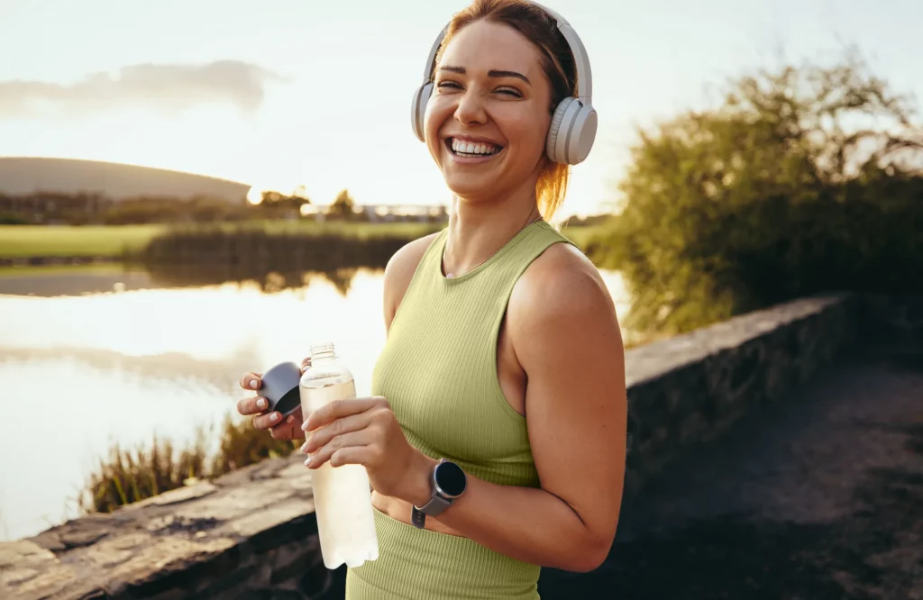 Eine Frau mit Kopfhörern joggt und macht in der Natur eine Trinkpause.