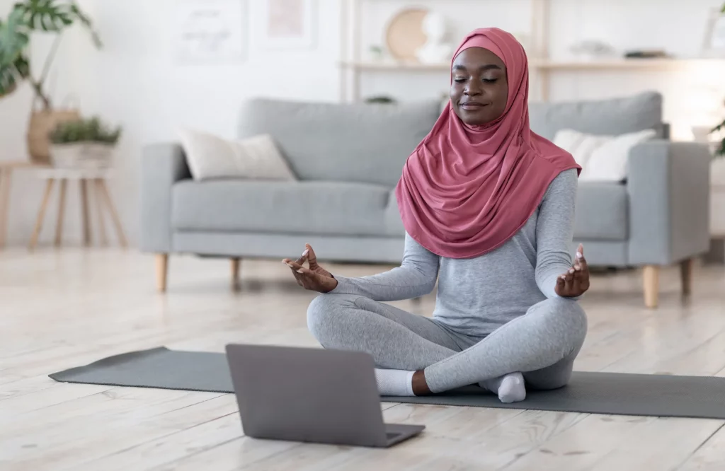 Eine Frau mit Kopftuch meditiert zu Hause vor ihrem Laptop während einer Online-Yoga-Stunde.