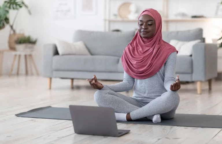 Eine Frau mit Kopftuch meditiert zu Hause vor ihrem Laptop während einer Online-Yoga-Stunde.
