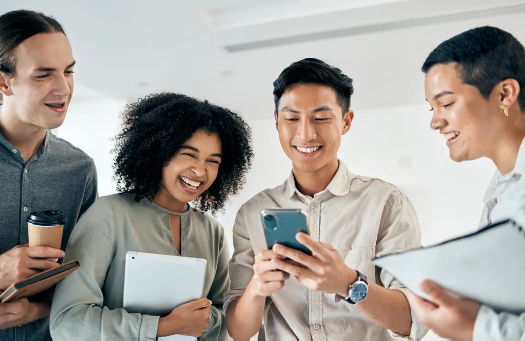 Eine Gruppe von Kollegen steht lachend zusammen und betrachtet ein Smartphone während einer Pause im Büro.