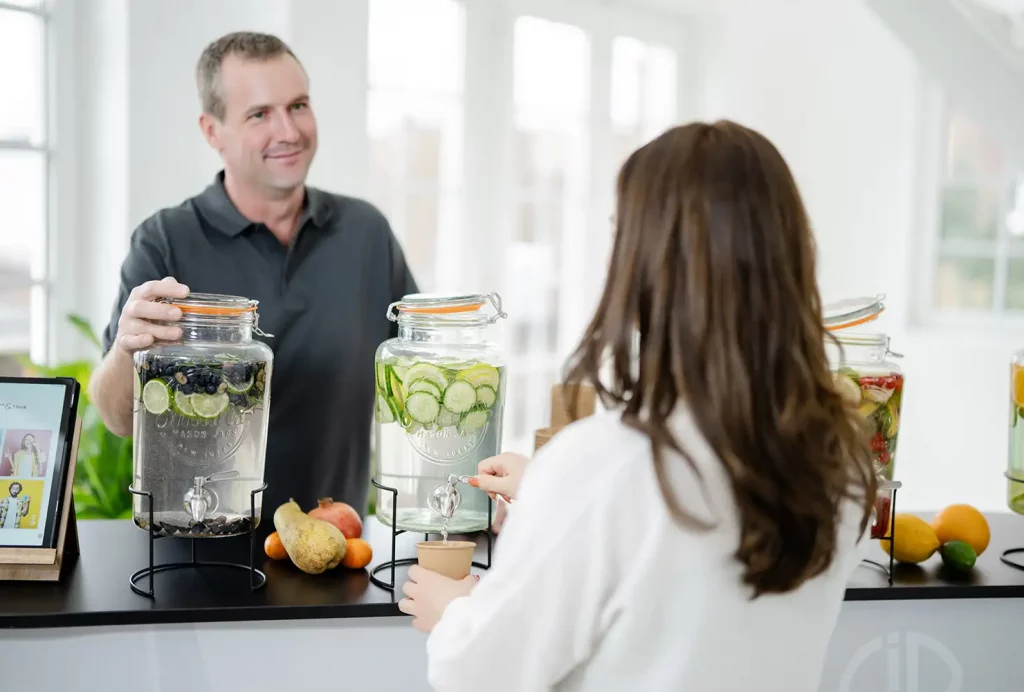 Ein GiB-Trainer berät eine Teilnehmerin, während sie sich frisches Infused Water mit Gurke und Zitrone am Getränkespender einschenkt.