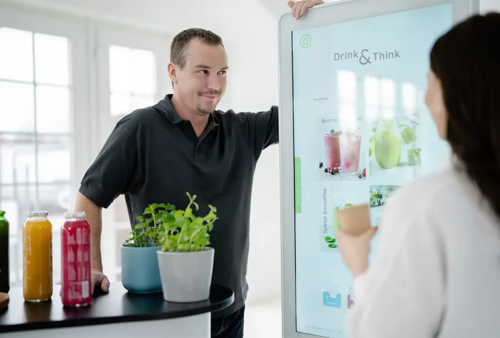 Nahaufnahme eines Beratungsgesprächs: GiB-Trainer und Teilnehmerin mit einer Smoothie-Probe an der interaktiven Touch-Stele „Drink & Think“.