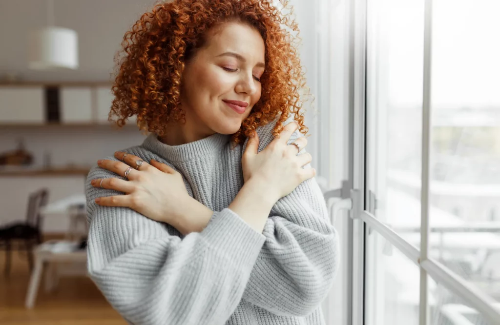 Frau ist zufrieden mit sich und umarmt sich selbst am Fenster.