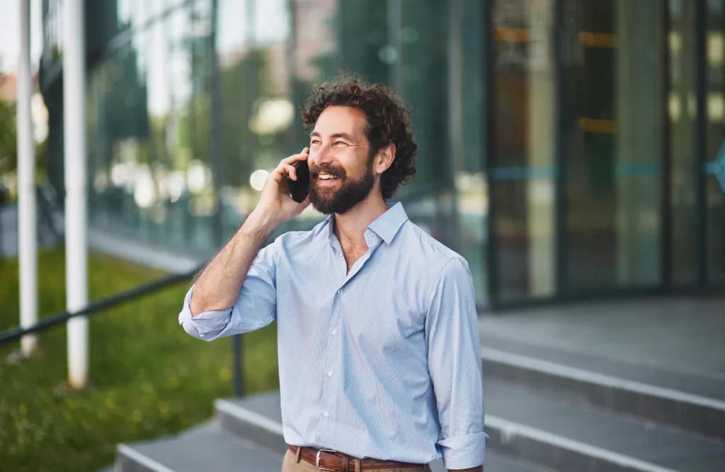 Ein lächelnder Geschäftsmann telefoniert im Gehen mit einem Smartphone vor einem modernen Bürogebäude.