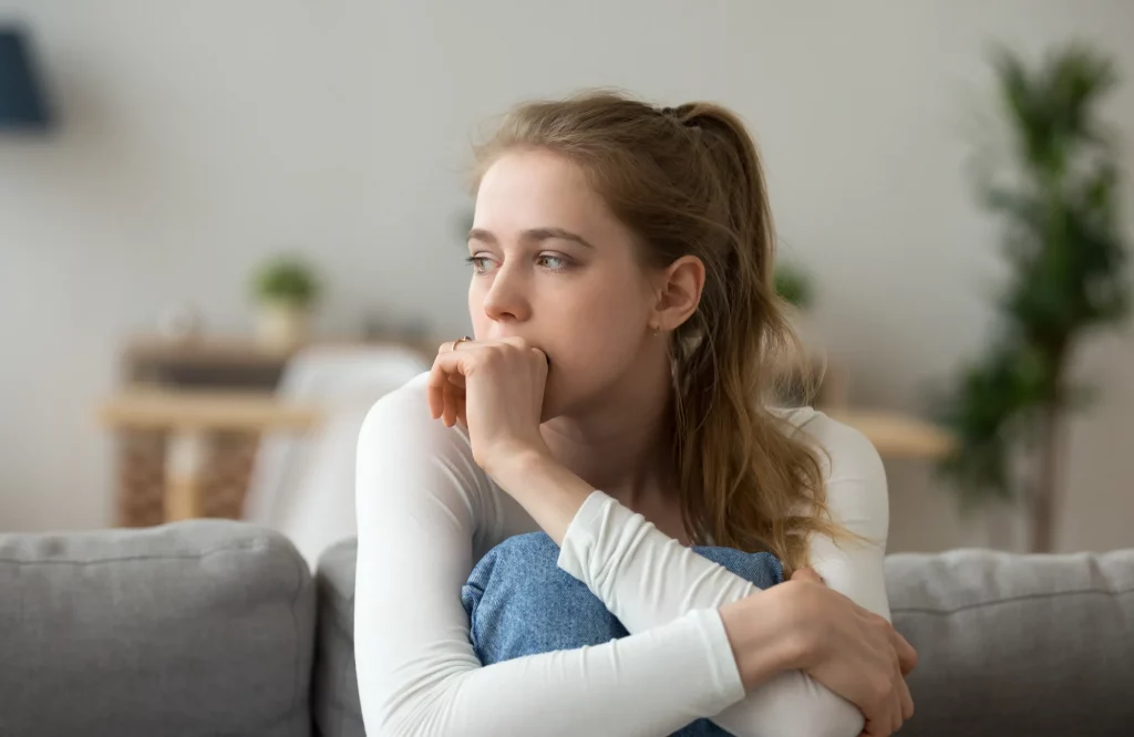 Eine besorgte Frau sitzt mit nachdenklichem Blick auf ihrem Sofa zu Hause.