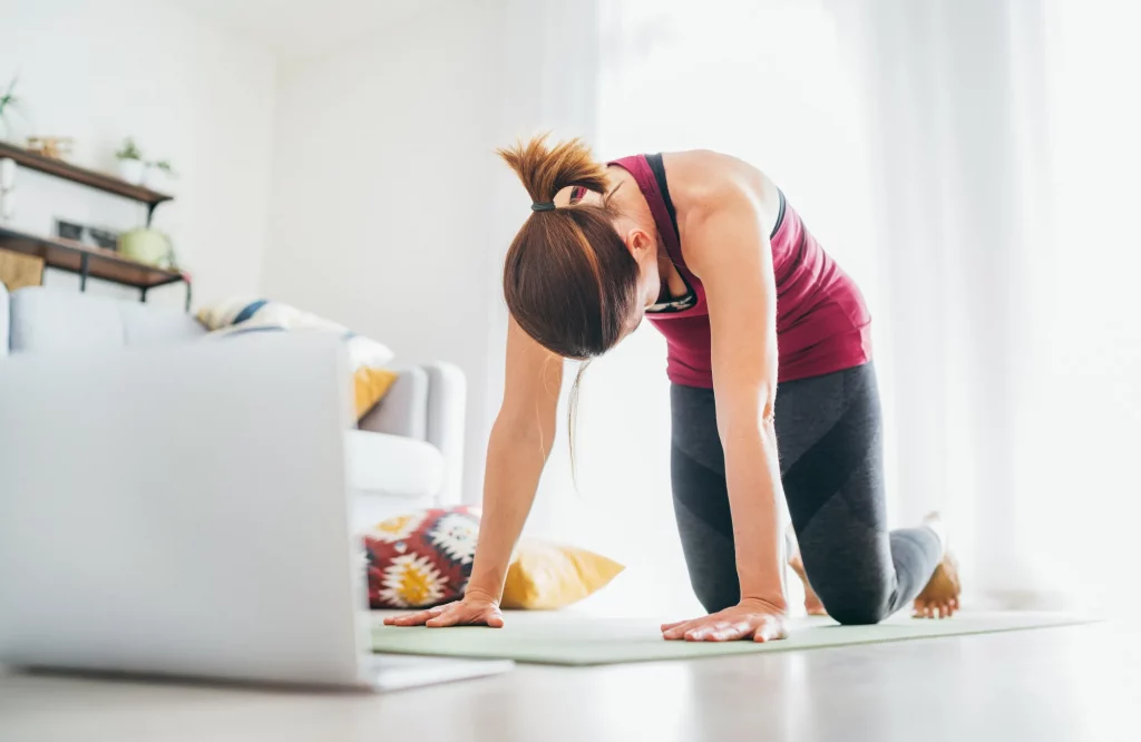 Eine sportliche Frau in der Yoga-Position „Katze“ (Majaryasana) macht Atemübungen vor ihrem Laptop.