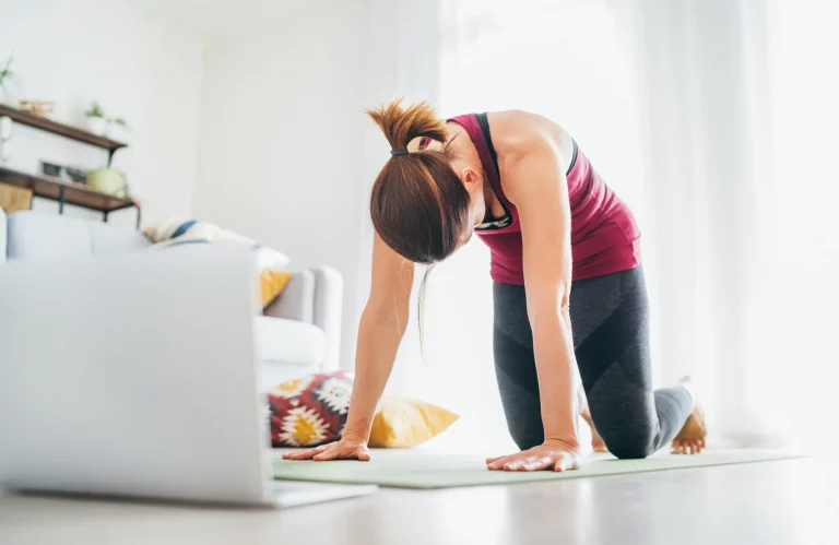 Eine sportliche Frau in der Yoga-Position „Katze“ (Majaryasana) macht Atemübungen vor ihrem Laptop.