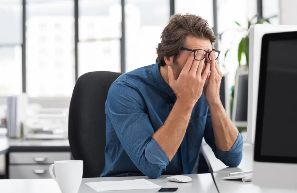 Geschäftsmann sitzt gestresst am Schreibtisch und reibt sich müde die Augen bei der Arbeit im Büro.