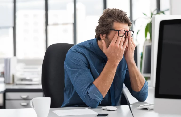 Geschäftsmann sitzt gestresst am Schreibtisch und reibt sich müde die Augen bei der Arbeit im Büro.