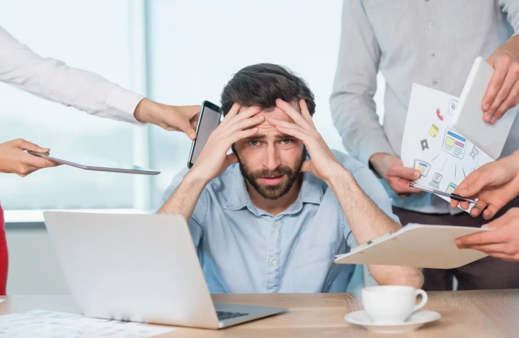 Gestresster Mann im Büro schlägt die Arme über dem Kopf zusammen, während Kollegen ihm neue Aufgaben via Telefon und Dokumenten überreichen.