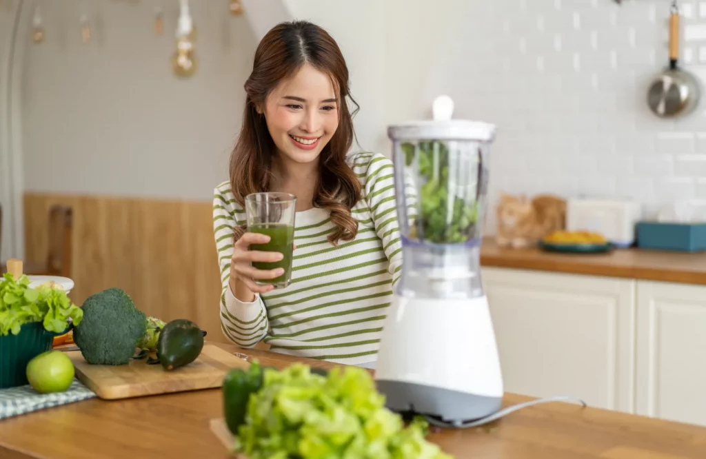 Eine Frau bereitet in der Küche einen grünen Smoothie mit einem Mixer zu und trinkt aus einem Glas.