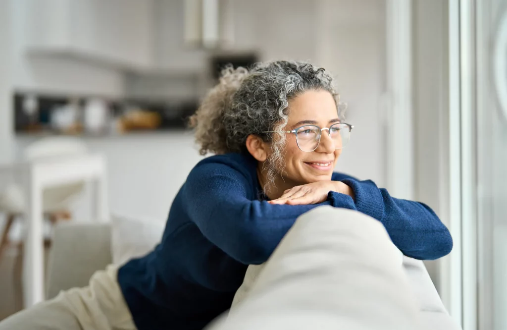 Eine Frau mit Brille sitzt entspannt auf einem gemütlichen Sofa und blickt lächelnd aus dem Fenster.