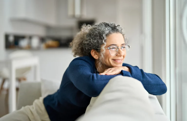 Eine Frau mit Brille sitzt entspannt auf einem gemütlichen Sofa und blickt lächelnd aus dem Fenster.
